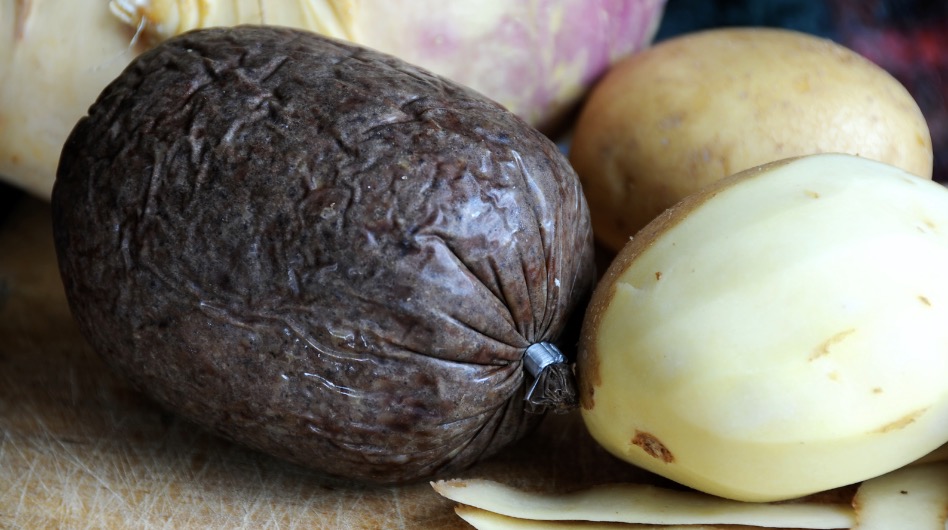 A dark roll of haggis in the center left of the image. Beside it are two half-peeled potatoes. The haggis is still within its outer casing.