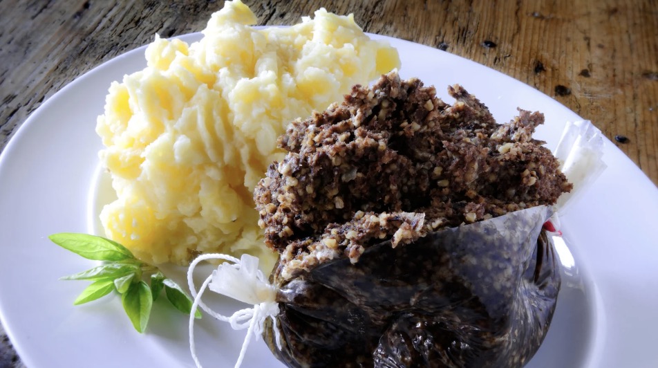 A plate of haggis and mashed potatoes. The haggis is half in its outer casing, but mostly coming out of it. The haggis is a stuffing-like texture to make up the traditional Scottish pudding. It is dark brown with some reddish and tan variation. The mashed potatoes above it are yellow and creamy. Beside is a small amount of basil.