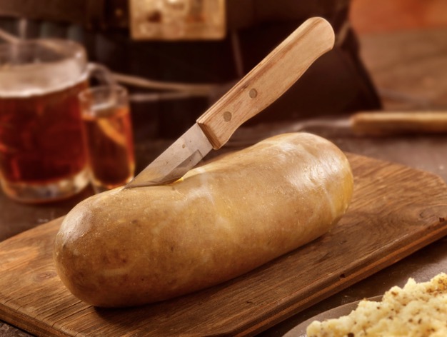 A light roll of haggis sitting on a wooden cutting board. Behind it is a pitch of amber liquid and a glass. The haggis has a knife protruding out of the top, stuck in the moment of slicing open the haggis.
