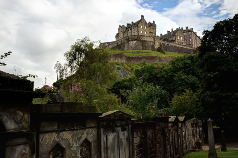 A lush scene of the Scottish Highlands. An old wall with ornate carvings runs along the bottom of the hill. A multitude of different trees grow in the middle ground. Atop the hill is a large tan and gray castle. A cloudy and blue sky sets the scene.