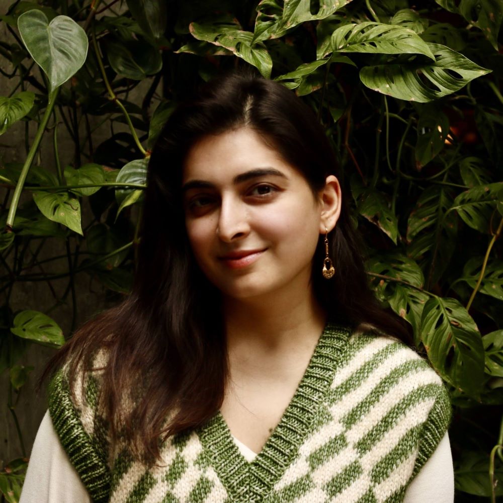 Maham Khwaja, a smiling brown-skinned woman with long, dark, straight hair wearing a green-and-cream patterned sweater vest in front of a wall of climbing leaves.