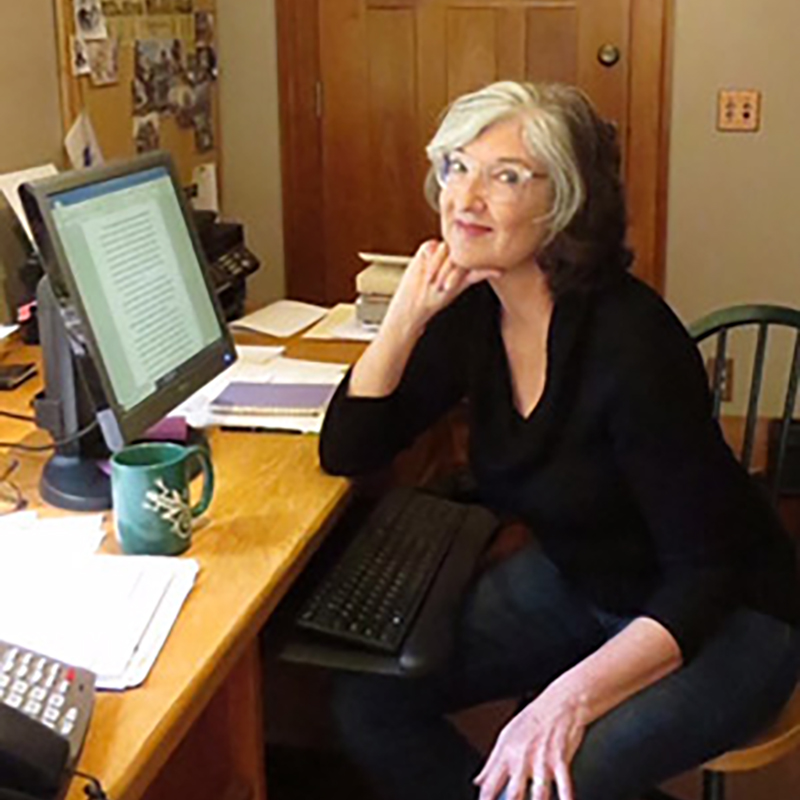 Barbara Kingsolver wears a black shirt, jeans, and clear glasses. She sits at a desk with her chin resting on her fist. A computer with an open document on the screen is in front of her. The desk top is covered with papers, books, a printer, a phone, and a green mug. Behind is a wooden door and a corkboard covered with pictures.