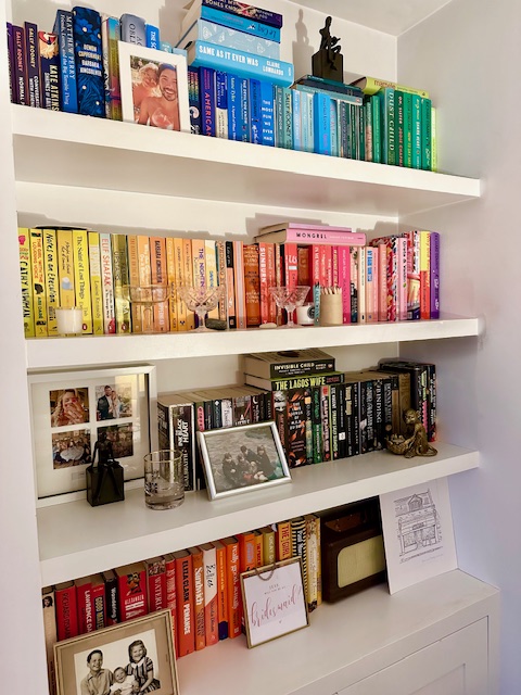 A white bookshelf with books organized by color. The top shelf has blue and green books. The second shelf has yellow and pink books and decorative glasses. The third shelf has black books, framed photos, a glass, and two figurines. The bottom shelf has red and orange books and a sketch of a building.
