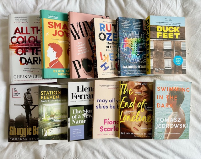 White bedsheets. On top are two rows of multicolored books. There are twelve books in total. Only some titles can be seen because the covers are overlapping each other.