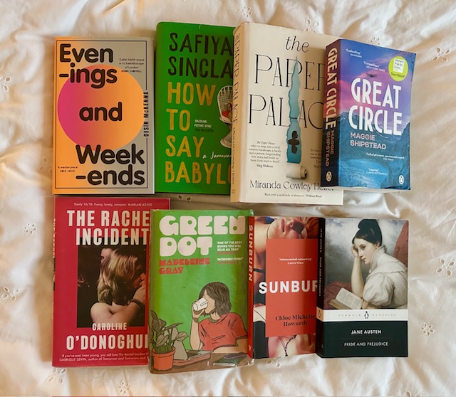White bedsheets. On top are tow rows of books. There are eight books in total. Titles are: Evenings and Weekends, How to Say Babylon, The Paper Palace, Great Circle, The Rachel Incident, Green Dot, Sunburnt, and Pride and Prejudice.