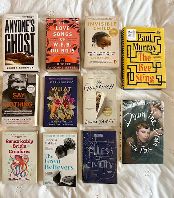 White bedsheets. On top are 11 books: Anyone's Ghost, The Love Songs of W.E.B. Du Bois, Invisible Child, The Bee Sting, Say Nothing, What My Bones Know, The Goldfinch, Down the Drain, Remarkably Bright Creatures, The Great Believers, and Rules of Civility.