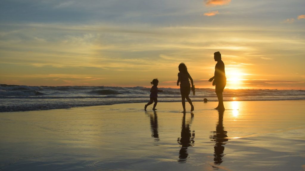 A family of three — including a mother, father, and young daughter — run along the edge of the ocean on a beach as the sun sets behind them, casting the sky in shades of blue and orange.