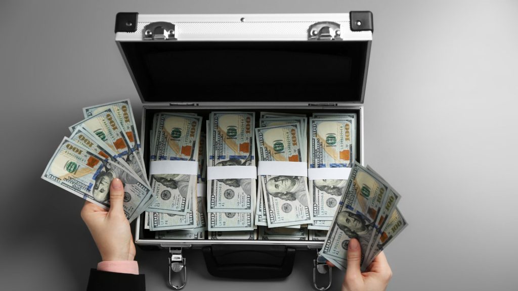 A metal briefcase filled with bundles of $100 bills. Two hands hovering above the briefcase hold stacks of bills.