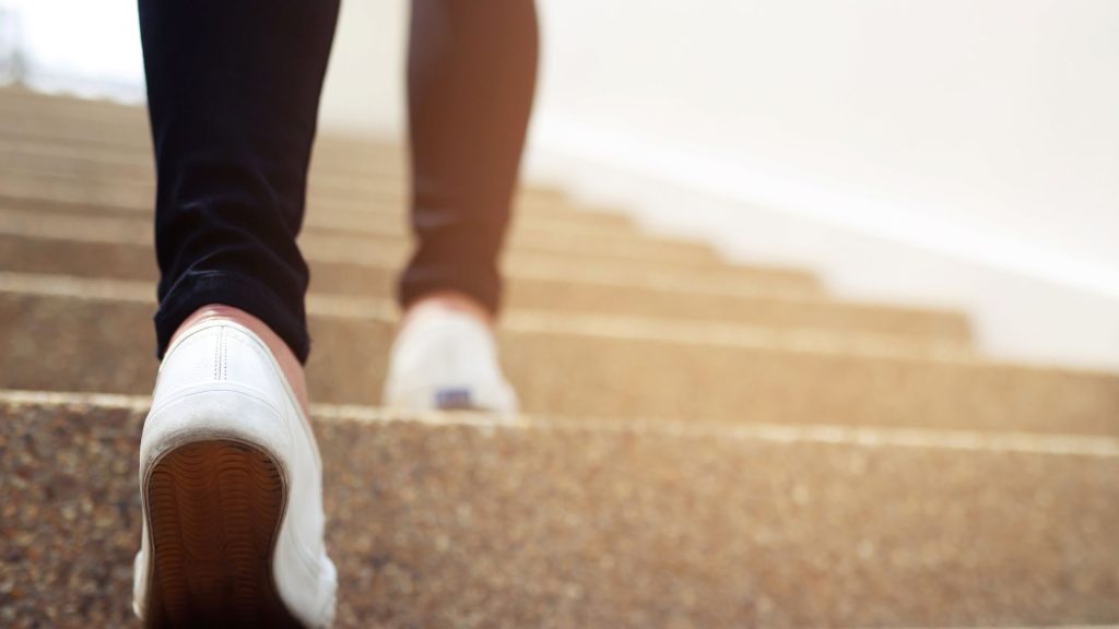 A person wearing black pants and white sneakers walks up a flight of concrete stairs.