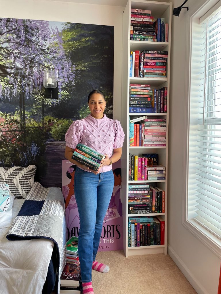 Krystal Marquis wears blue jeans, a short sleeve purple sweater, and pink socks. She stands in front of a blossom tree mural painting, a tall bookshelf, and a bed. She smiles and holds a stack of books in her hands.