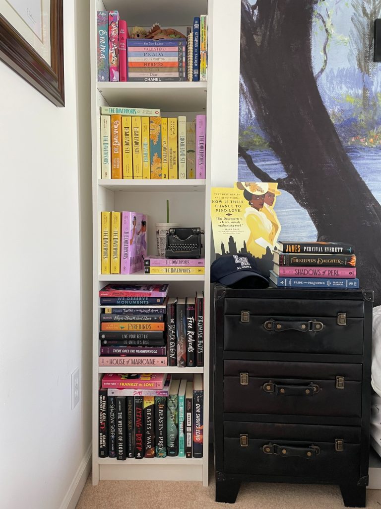 A tall white bookshelf full of books. Beside it is a short black nightstand. On the top of it is a stack of books, a blue baseball cap, and a small yellow poster for The Davenports.