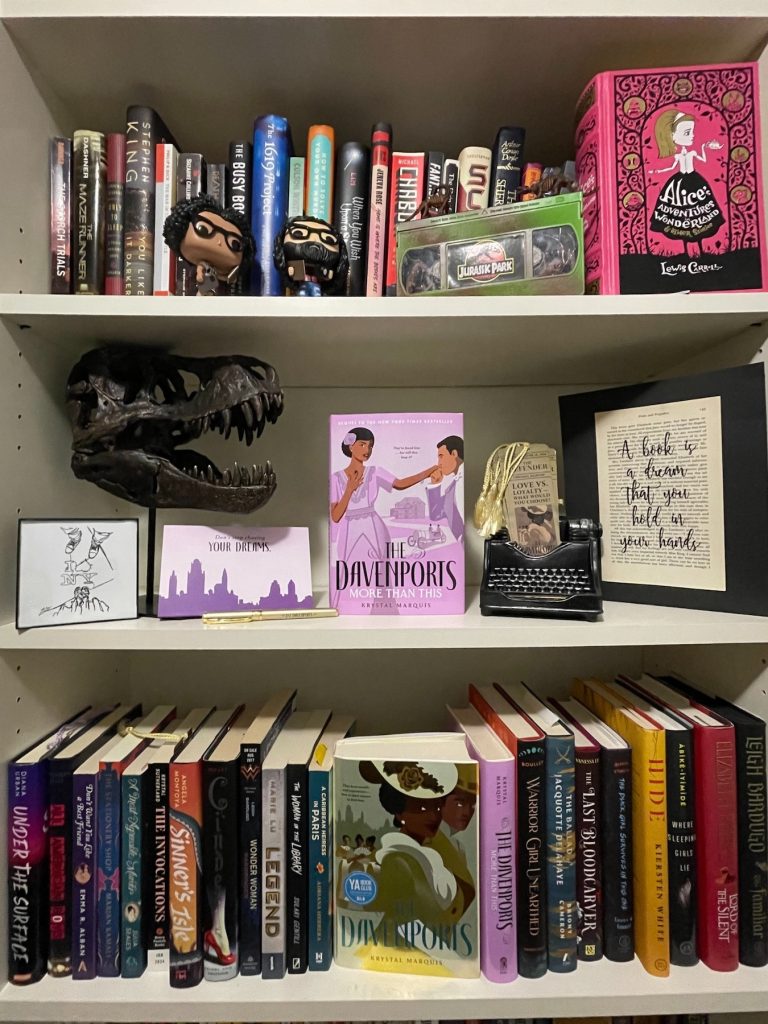 Three bookshelves full of books, Jurassic Park memorabilia, two Funko Pop figurines, a small novelty typewriter, and mounted quote art.