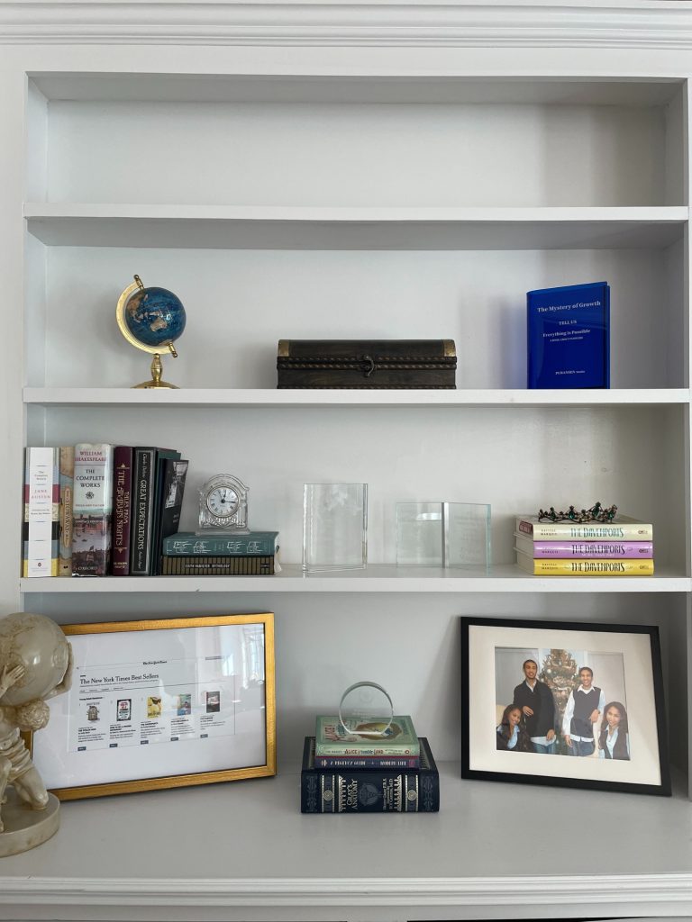A white bookshelf with some books, awards, a small globe, a marbled statue, a clock, and framed photos.