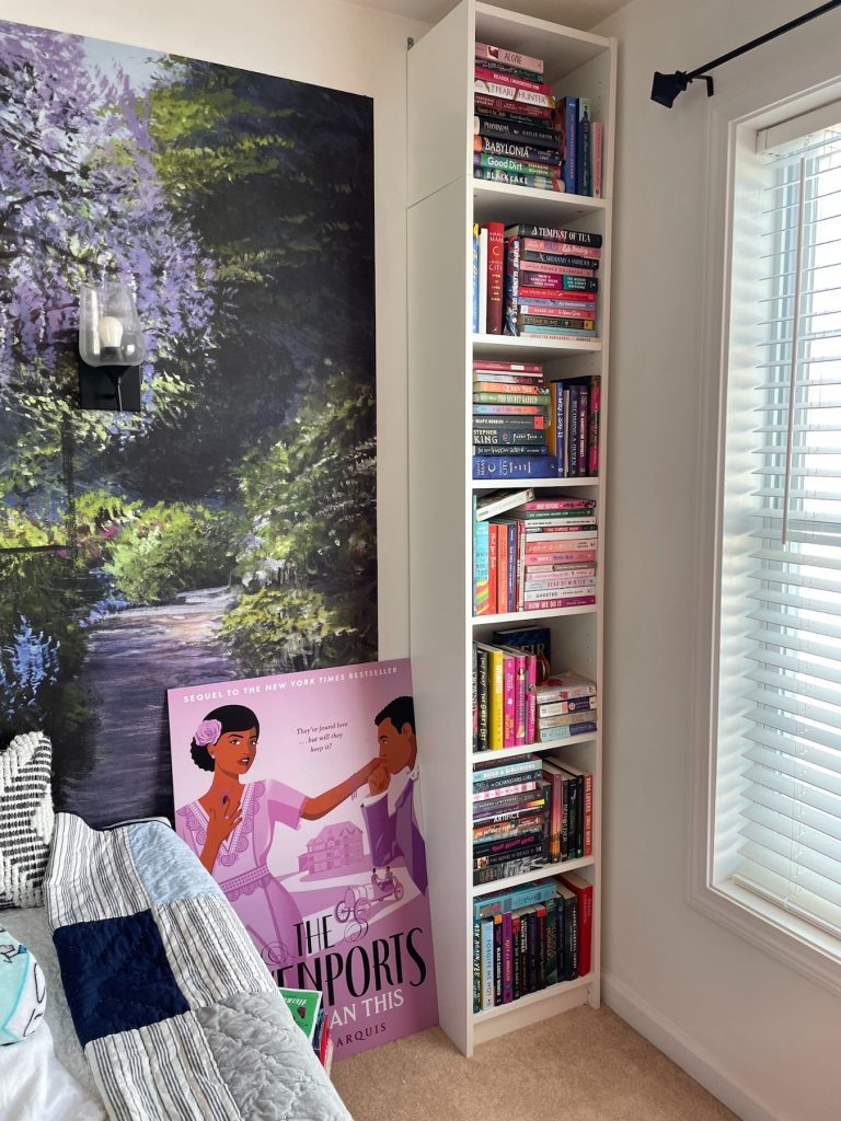 A tall white bookshelf full of books. Beside it is a floral mural painting and a purple poster of The Davenports leaning against the wall.