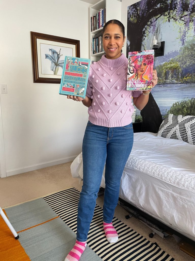 Krystal Marquis wears blue jeans, a short sleeve purple sweater, and pink striped socks. She stands in front of a bed with white sheets and holds two special edition books in her hand. One is Alice's Adventures in Wonderland and the other is Pride and Prejudice.