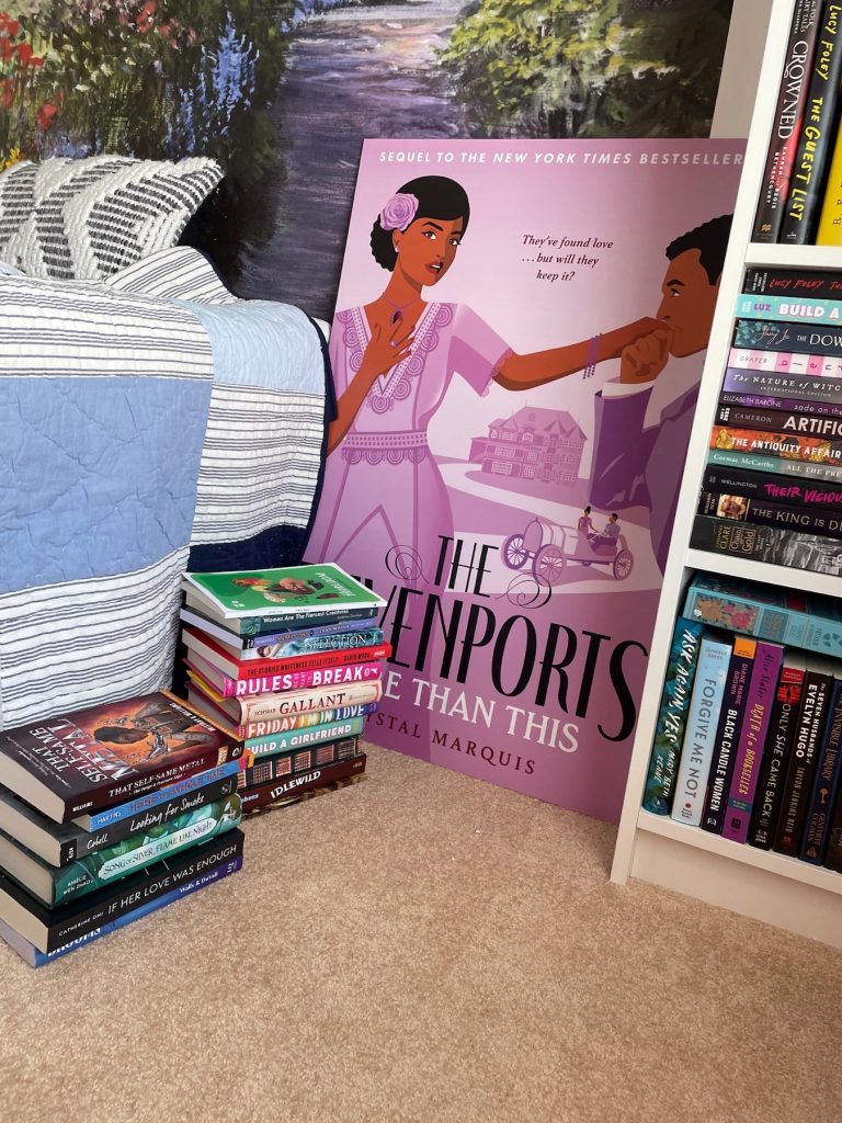 The floor beside a full white bookshelf. Two stacks of books beside a bed with gray and white striped sheets. A purple poster for The Davenports: More Than This leans against the a floral mural painting on the wall.
