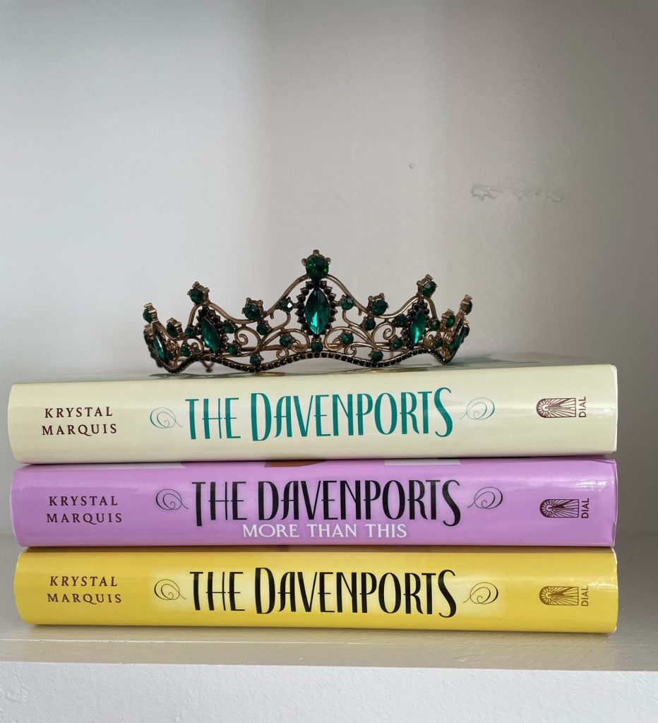 Three editions of The Davenports stacked on top of a white bookshelf. The top cover is off-white with teal lettering. The middle cover is purple with black lettering. The bottom cover is yellow with black lettering. A tiara with green jewels rests on top of them.