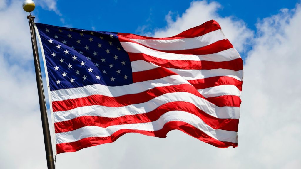An American flag on a flagpole waving in front of a cloudy blue sky.