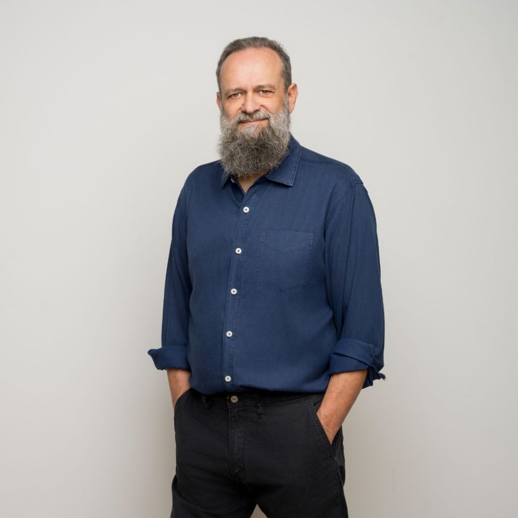 Author Mir Bahmanyar wears a blue button-down shirt and black pants. He has a long gray beard and stands in front of a gray background with his hands in his pockets, slightly smiling.