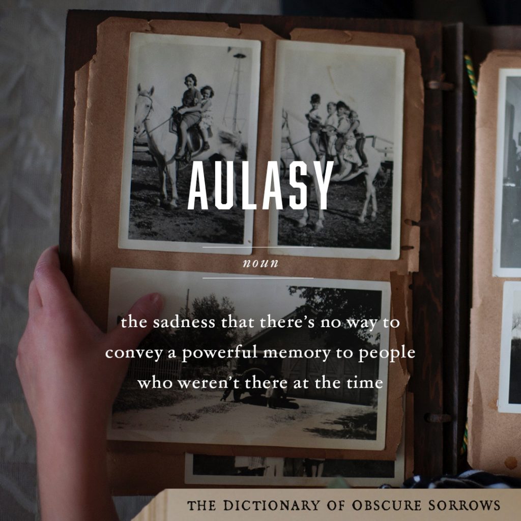 A hand holds open a brown album full of black and white photographs. White text on top reads: aulasy, noun, the sadness that there's no way to convey a powerful memory to people who weren't there at the time. In the bottom right corner is a light brown banner that says The Dictionary of Obscure Sorrows in black lettering.