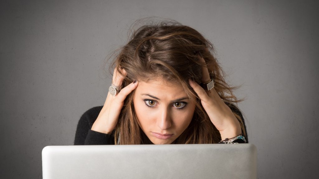 A gray background. A woman sits in front of a laptop and looks stressed. Her hands are in her hair. She wears a black shirt, two large silver rings, and a brown and teal bracelet.