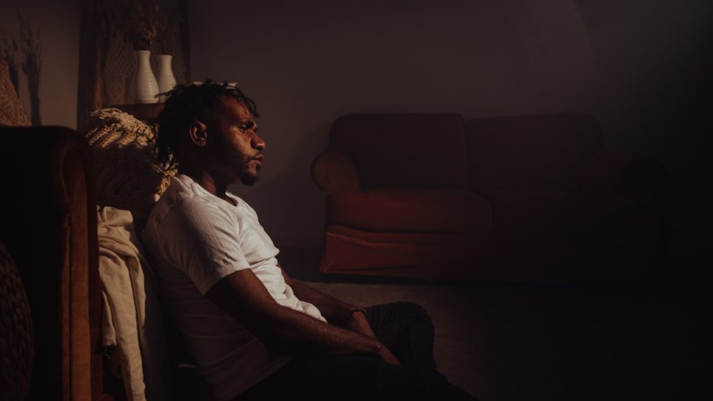 A man wearing a white shirt sits on the floor against a brown couch and looks pensive. There is another brown couch, book shelves, and vases in the background. The room is dimly lit.
