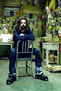 Rob Zombie posing on the set of Halloween (2007). He is sitting on a reversed metal chair, wearing dark jeans and a long-sleeved flannel. In the background, the walls are covered in different horror-themed masks. There are also creepy worktables behind him.