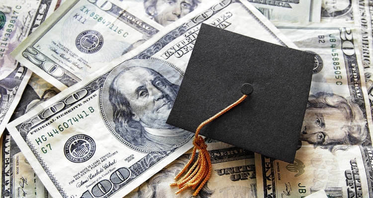 Image shows multiple pieces of cash covering the background as a graduation cap sits on top of them.