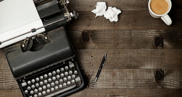 Image shows a typewriter on the left side and a cup of coffee in the top right corner with a pen and crumpled papers in the middle of the desk.