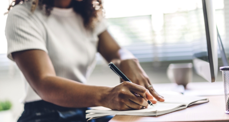 Image shows a woman writing in a journal on a desk. Their desktop sits on the far right side.