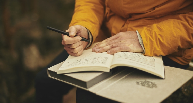 Image shows someone writing in a journal on top of a book sitting in their lap.
