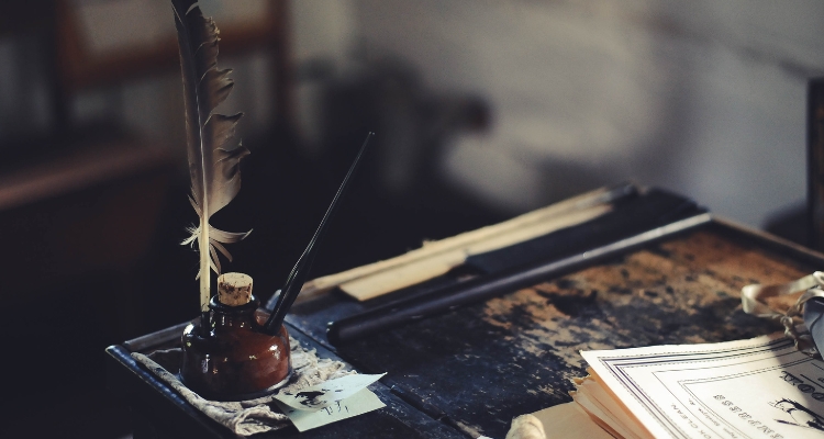 Image shows the corner of a desk along with the corners of papers and a quill next to an ink bottle.