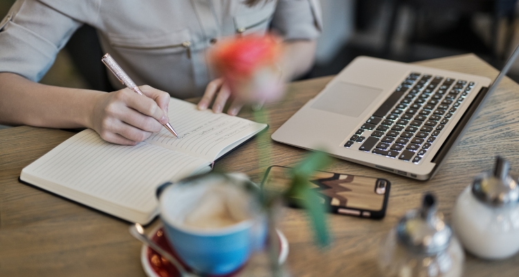 Image is of someone writing in a notebook on a desk with their computer in front of them. The view is slightly obscured by a blurry rose and cup of coffee.
