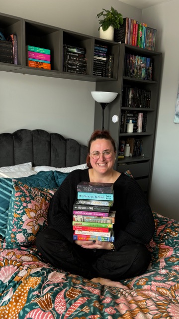 Cait sitting on a bed with a pink and green floral comforter holding a stack of colorful books. There are additional books behind the bed on a gray bookshelf.