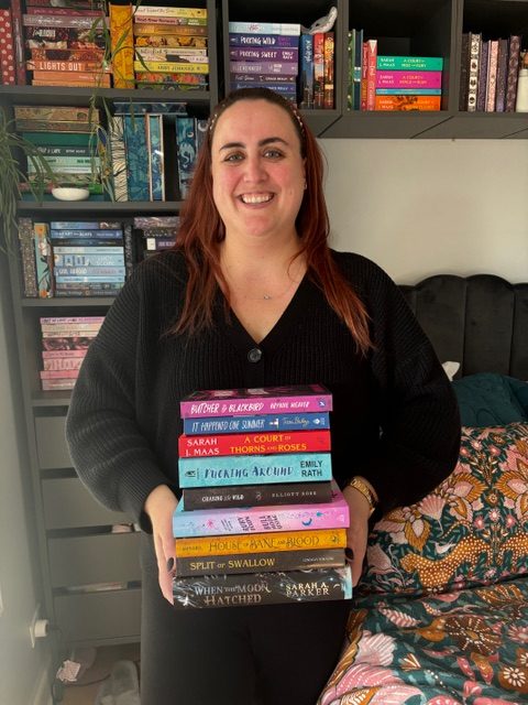 Cait pictured smiling in front of a full bookshelf holding a colorful stack of books while wearing a black sweater.