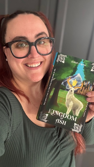 A selfie of Cait wearing a green long sleeve top and wide-rim glasses holding Kingdom of Ash by Sarah J. Maas.