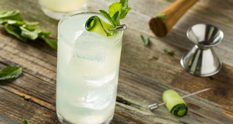 A light green drink in a tall glass with three large ice cubes and a rolled cucumber and mint garnish on a wooden table.