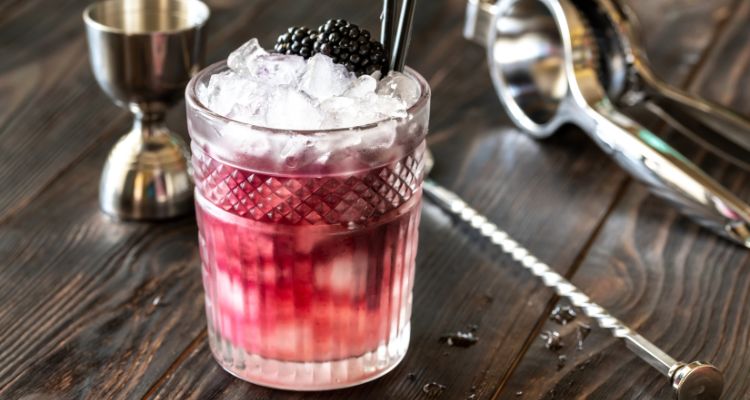 A light purplish pink drink sits on a wooden table in an etched crystal glass with ice on top and blackberries.