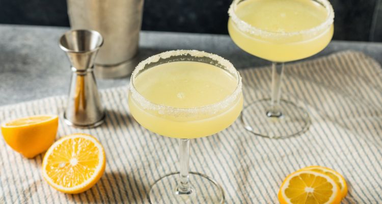 Two lemon gimlets in rounded glasses have a sugar rim and sit on a rustic green and beige pinstriped towel with lemons and lemon slices surrounding them.