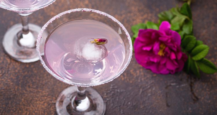 A martini glass with a sugar and rose petal rim holds a light pink liquid and there is a bright pink rose in the background.