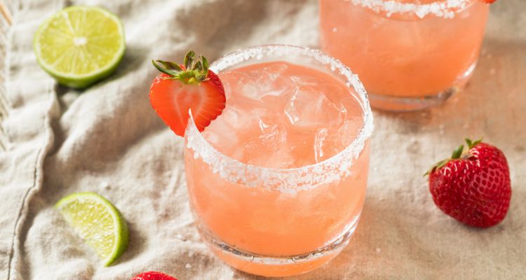 Two pinkish margaritas with a salt rim sit on a brown cloth towel with lime slices and a strawberry around them. The closer drink has a sliced strawberry on the rim.
