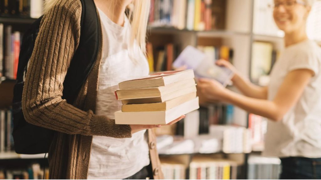 Two figures are at a bookstore, the figure closest to the camera is holding a stack of 5 books and wearing a white shirt with a brown cardigan and a backpack. The other figure is wearing a white shirt and jeans, holding a book.