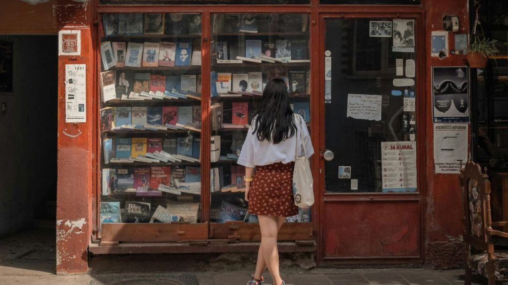 A girl with her back to the viewer is facing a wall of books displayed on a shelf inside a bookstore. She is wearing a red designed skirt and a white shirt, holding a tote bag on her shoulder and has black mid-length hair.