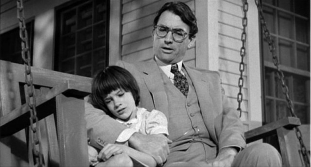 Black-and-white image from To Kill a Mockingbird film adaptation from Universal Pictures depicting a father and daughter embracing another while sat on a bench.