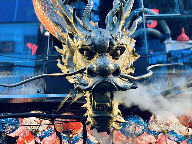 A close-up of a golden Chinese dragon sculpture with intricate detailing, fierce eyes, and whiskers extending from its snout. Steam emerges from its mouth, adding a dramatic effect. The background features colorful paper lanterns and glass windows reflecting city lights.