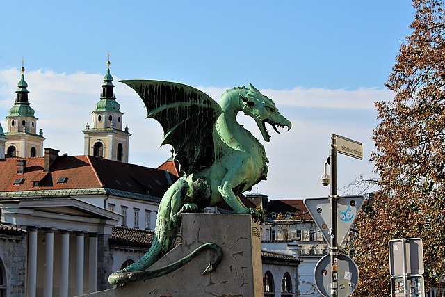 The iconic green dragon statue atop the Dragon Bridge in Ljubljana, Slovenia, with historic European architecture and church towers in the background. The bridge is a famous landmark and symbol of the city.
