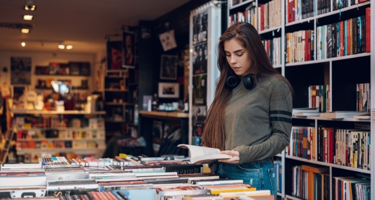 A woman with headphones around her neck looks at a book inside a bookstore.