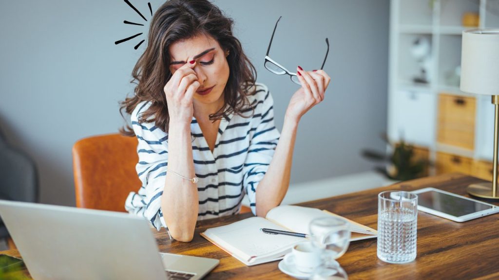 Woman sitting at a wooden table with a laptop, planner, and cups of coffee and water, holding her glasses with one hand and pinching her nose with the other, showing signs of stress and tiredness.