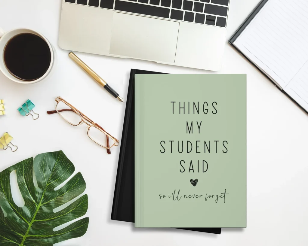 A green journal with the title, "Things My Students Said So I'll Never Forget," on a white desk surrounded by a pair of glasses, a pen, a laptop, a notebook, a plant leaf, and a coffee.
