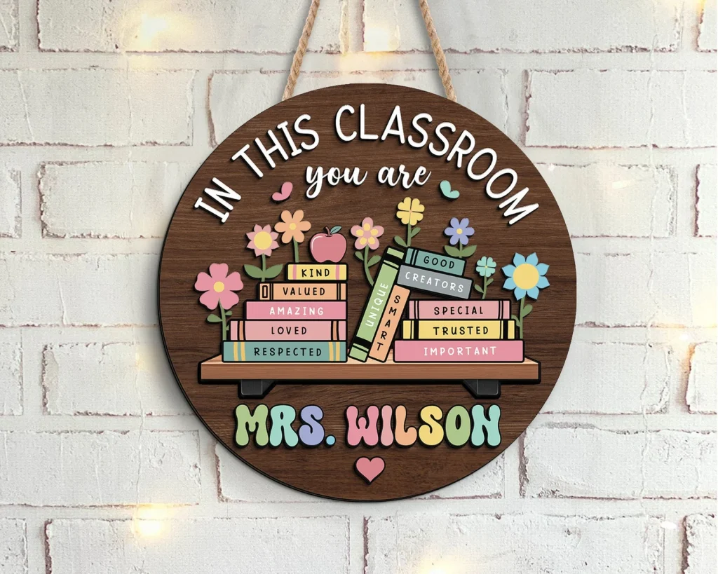 A round brown piece of wood with a rope to make it a door hanger. There are white words at the top of the circle that says, "In this classroom you are" and then there are colorful books underneath that tells the reader what they are. The books are titled; kind, valued, amazing, loved, respected, unique, smart, good, creators, special, trusted, and important. There are flowers sprouting out from the books, an apple on top of the "kind" book, and two butterflies flying over the flowers. Underneath the stack of books is the teacher's name. This example uses the name Mrs. Wilson spelled out with different colored letters.